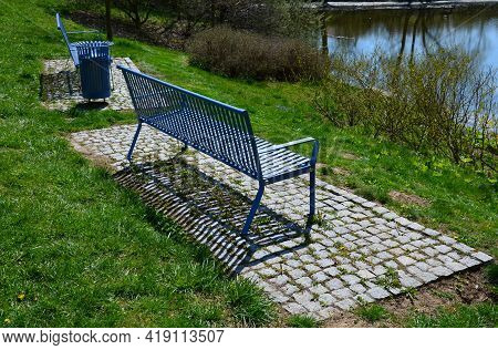 Park Benches In Light Blue Made Of Metal Strips Similar To A Lattice. Trash Can And Beautiful Lawn W