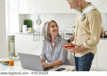 Senior Husband Preparing Breakfast To Happy Wife Working From Home Office Using Laptop Sitting At Ki
