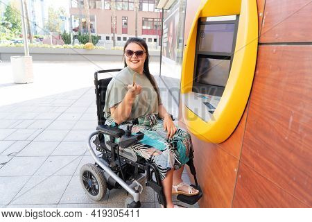 Young Happy Disabled Handicapped Woman In Wheelchair Using Atm Cash Macjine And Smiling At Camera On