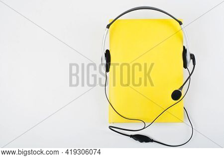 Audiobook On White Background. Headphones Put Over Yellow Hardback Book, Empty Cover, Copy Space For