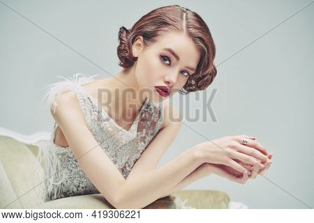 Portrait of a beautiful chic woman posing in a luxury white dress in vintage armchair. Evening makeup and hairstyle of the 20s. 