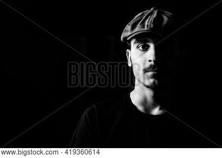 Black And White Portrait Of A Young Guy With Moustache And A Hat On Black, Dark Background.