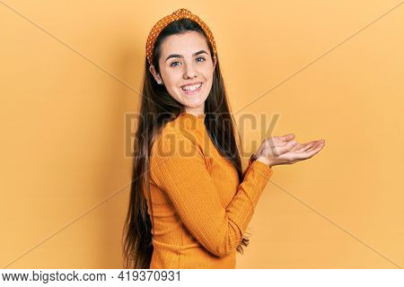 Young brunette teenager wearing casual yellow sweater pointing aside with hands open palms showing copy space, presenting advertisement smiling excited happy 