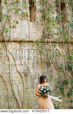 Beautiful Bride In A Cream Wedding Dress With A Bouquet Of Flowers On The Background Of A Stone Wall