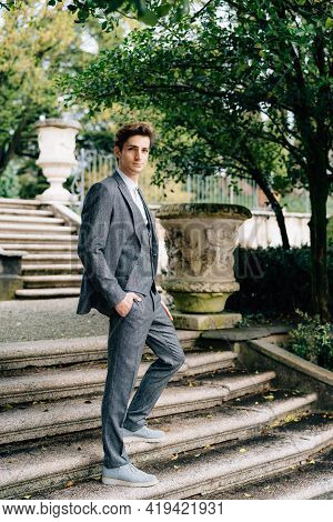 Serious Groom Stands With His Hands In His Pockets On The Stairs In The Park Near The Old Villa. Lak