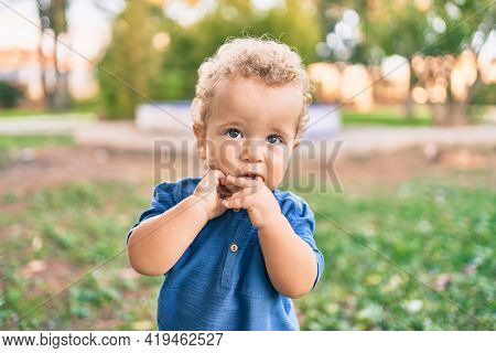 Sad little boy putting fingers on mouth touching gums because toothache at the park on a sunny day. Beautiful blonde hair male toddler in pain for new baby teeth outdoors