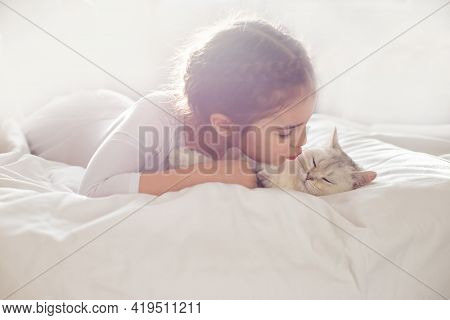 Portrait Of A Beautiful Little Girl, Lies On A Clean, White Bed With A White British Cat.