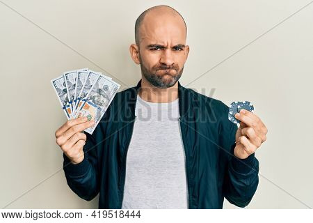 Young hispanic man playing poker holding dollars banknotes and chips skeptic and nervous, frowning upset because of problem. negative person. 