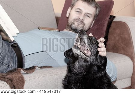 A Middle Aged Man With Greyish Hair And Beard Lying On The Sofa Nad Petting His Black Senior Dog