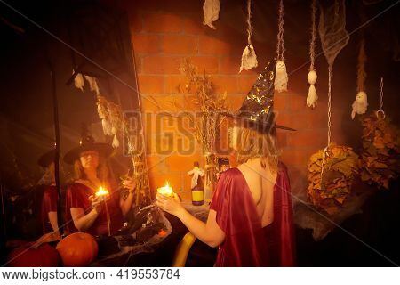 Blonde Witch In Red Dress And Black Hat With Candle And Mirror In Halloween Decoration Indoors.