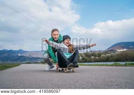 Laughing Kids Have Fun Using A Skateboard. Brother Pushing His Sister Sitting On A Skateboard And Ma