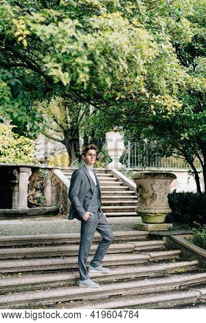 Serious Groom Stands With His Hand In His Pocket On The Stairs In The Park. Lake Como