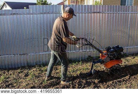 Male In The Spring Plows The Earth In Working Clothes With A Motoblock, Walk-behind Tractor