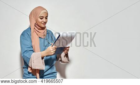 Happy Young Arabian Woman Doctor In Hijab Looking Into Notepad While Posing On Light Background With