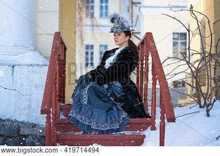 A Woman Dressed As A 19th-century Noblewoman Sits On A Porch Near The Castle