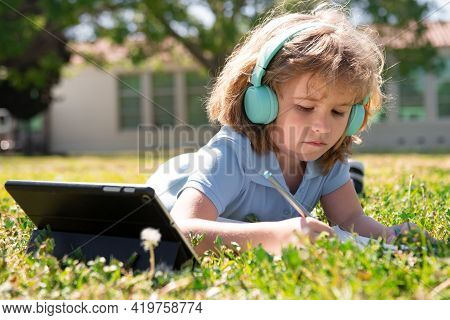 Child Homework Outside In Scholl Yard. Little Schoolboy Pupil With Tablet In The Park On Grass. Self