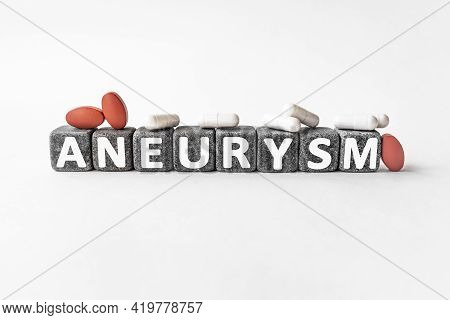 Aneurysm The Word On Stone Cubes. Cubes Stand On A White Surface, Many White And Red Pills. Medicine