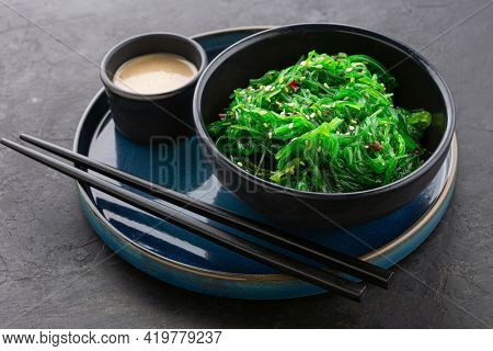 Healthy wakame seaweed salad in a black bowl, peanut sauce and chopsticks on a beautiful blue plate on a black slate background. Angle view.