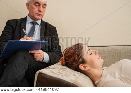 Caucasian Woman Lies On A Couch At A Reception With A Psychotherapist. An Elderly Man Works As A Psy