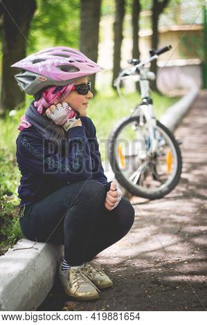 Konstantinovka, Ukraine - May 08, 2021: A Little Girl In A Helmet Sits On A White Curb. Warm Sunny S