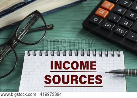 Income Sources - Words In A Notepad On A Wooden Green Background With A Calculator, Pen And Glasses.
