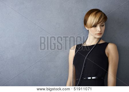 Elegant young woman in black dress leaning to wall, looking serious