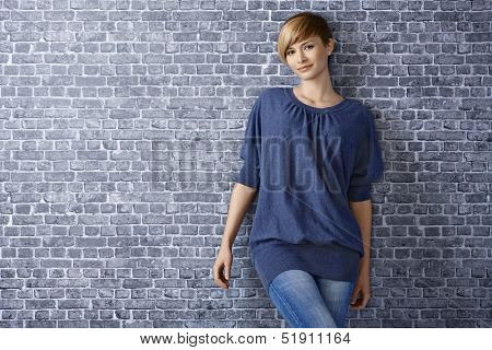 Portrait of casual young woman leaning to grey brick wall, smiling