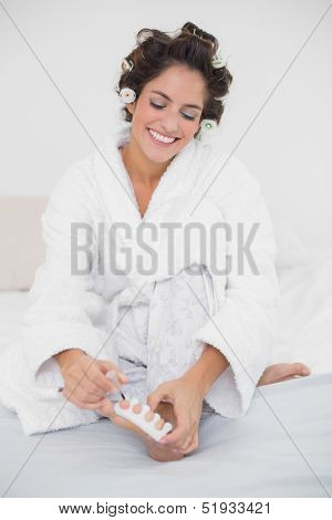 Happy natural brunette applying nail polish to her toenails in bedroom