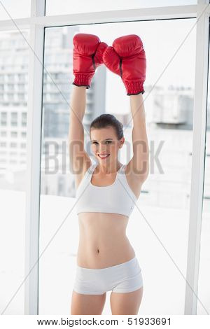Victorious fit brown haired model in sportswear wearing red boxing gloves in bright fitness studio