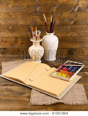 Composition with colorful watercolors, brushes in vase and sketcher on wooden background 