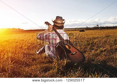 Girl In A Hat Sits On The Field And Holds The Guitar Behind His Back.