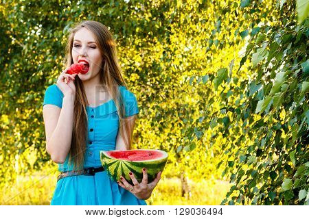 Beautiful blonde girl in a blue dress with long hair eating a watermelon outdoors