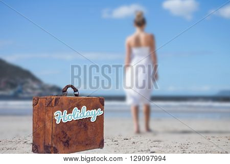 Blonde in white dress walking on the beach against suitcase with the message holidays