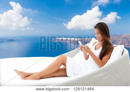 Asian woman using phone app relaxing on daybed on outdoor terrace. Home living outside patio furniture. Young adult texting on smartphone lying on sofa with Oia Santorini background enjoying summer. 