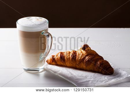 Glass of latte macchiato and French croissant on baking paper. Horizontal scene on white wooden background.