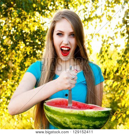 Beautiful blonde girl in a blue dress with long hair holds a half of watermelon with knife