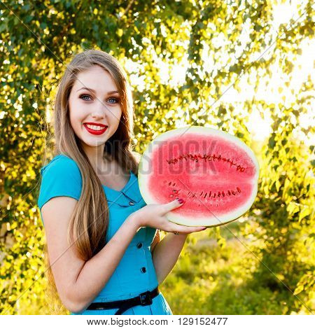 Beautiful blonde girl in a blue dress with long hair holds a half of watermelon backlight outdoors