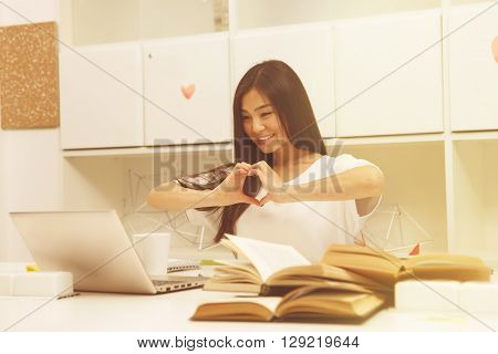 Toned image. Asian student communicating with friend abroad via Skype and showing heart while studying in library. Brunette lady smiling.