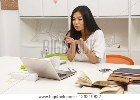Asian student lady doing air-kissing while communicating with foreign friend via Skype. Happy brunette studying in library.