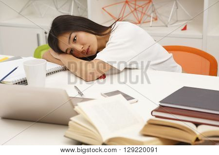 Tired Asian student resting after hard-working day in library. Exhausted woman lying on table with her eyes opened.