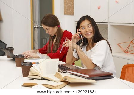 Happy Asian student lady studying and seaking over mobile or smart phone in library. Toothy smiling lady looking away.