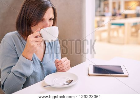 Beautiful mature caucasian woman taking a sip of her warm fresh cappaccino in a modern well lit coffee shop with her tablet lying on the table infront of her.