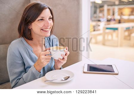 white woman with a beautiful smile busy smiling while looking off camera and holding a coffee as she looks off camera with her tablet lying on the table infront of her and excellent copy space.