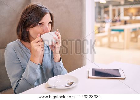 well dressed professional caucasian woman taking a sip of her coffee while sitting in a bright cafe with her tablet on the table infront of her.