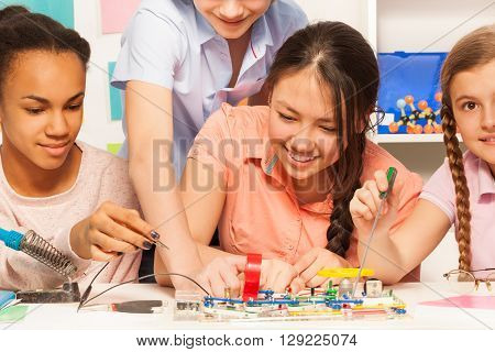 Four smiling pupils learning physics, assembling electric chain with electronic constructor