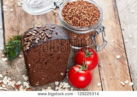 Close up photo of rustic bread sprinkled with sunflower seeds on sackcloth, two tomatoes, dill, oat flakes and jar with buckwheat on planked wooden table