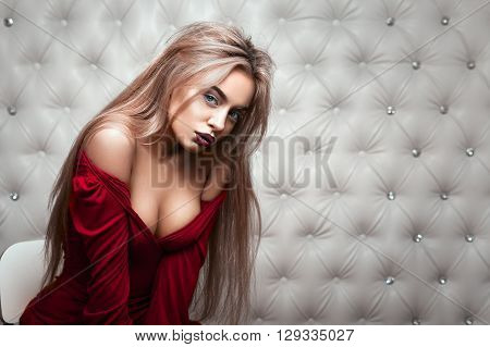 Studio portrait of a sexy blond in red dress, over leather upholstery background. Elegant luxury woman with a seductive neckline and bare shoulders looks into the camera.