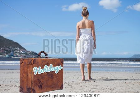 Blonde in white dress walking on the beach against suitcase with the message holidays