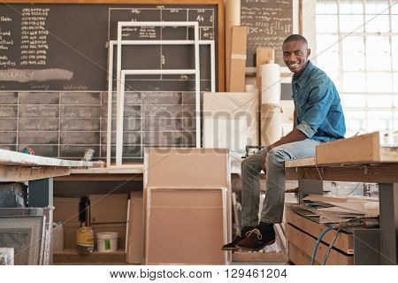 Portrait of a handsome young small business owner of African descent, smiling at the camera while sitting on the edge of a work bench in his studio workshop