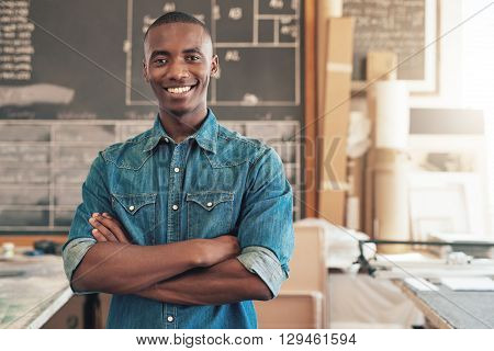 Portrait of a handsome young designer of African descent, smiling at the camera with his arms folded confidently, while standing in his studio